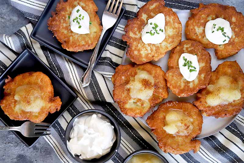 Horizontal overhead image of German potato pancakes on a white serving dish and two small, square black plates, with two forks, and small dishes of sour cream and applesauce, on a black, gray, and white striped cloth.