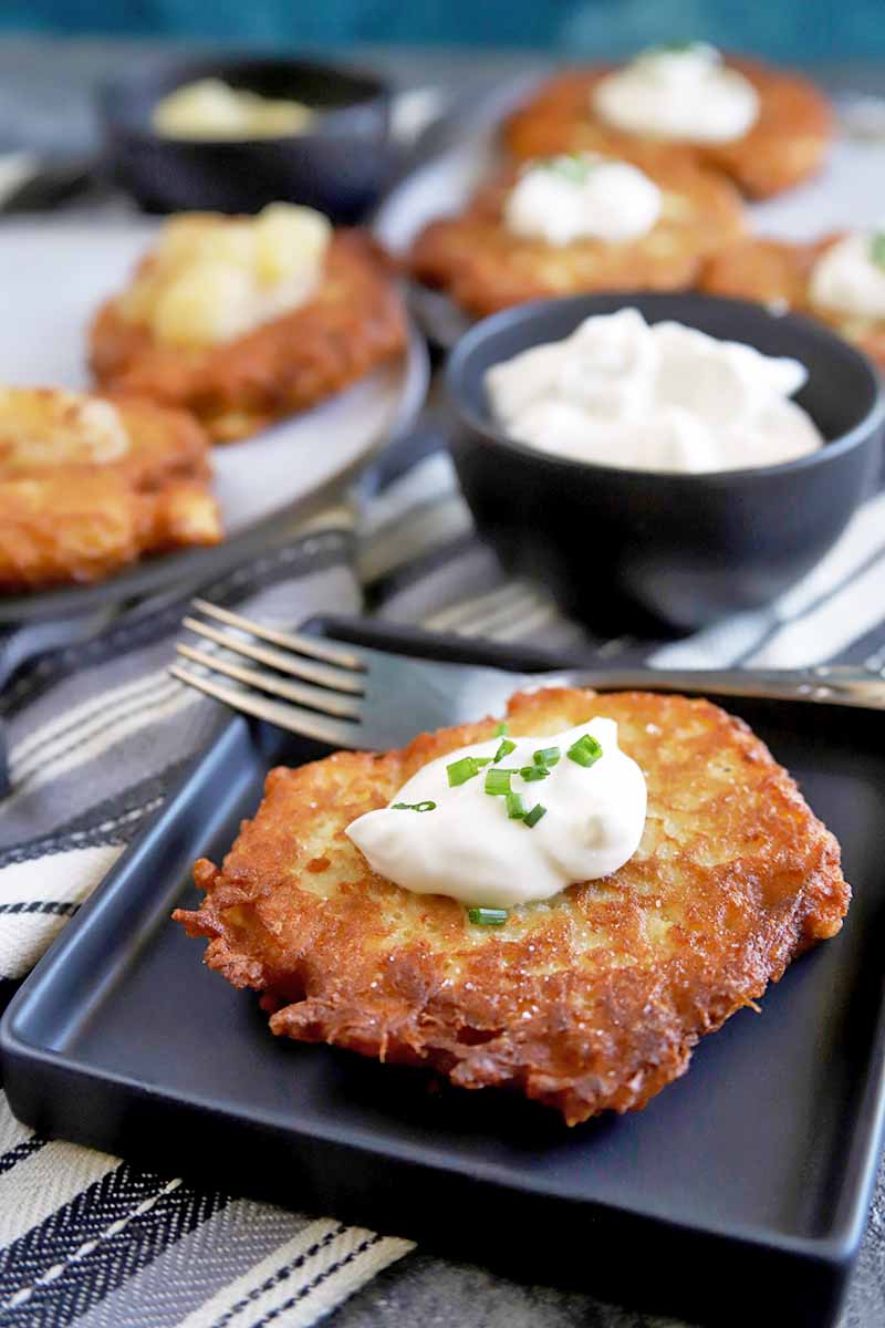 Vertical image of a German-style fried potato fritter topped with a dollop of sour cream and minced fresh chives, on a square black plate with a fork, with two small black bowls and two white serving plates containing more of the dish and the toppings in soft focus in the background, on a striped gray, white, and black cloth.