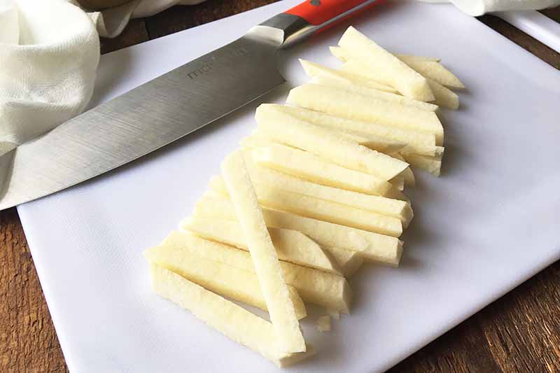 Horizontal image of a vegetable cut into French fry shapes on a white cutting board.