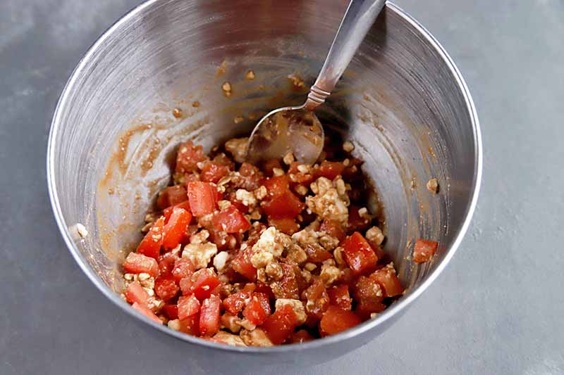 Horizontal overhead image of a stainless steel bowl containing a chopped tomato and crumbled feta mixture being mixed with a metal spoon, on a gray surface.