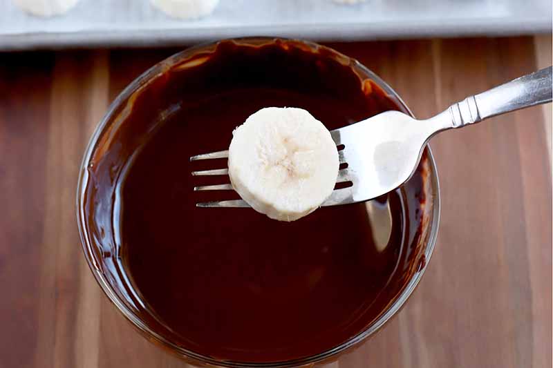 Horizontal image of a fork holding a banana slice over a small glass bowl of melted chocolate, on a wood surface with a baking sheet barely visible at the top of the frame.