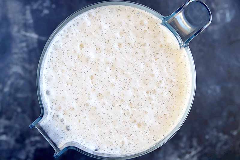 Overhead horizontal image of a frothy milk mixture in a plastic pitcher container, on a gray and white surface.