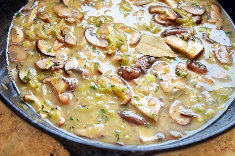 Horizontal image of a pan of mushrooms in sauce with a dried bay leaf on top, on a brown speckled surface.