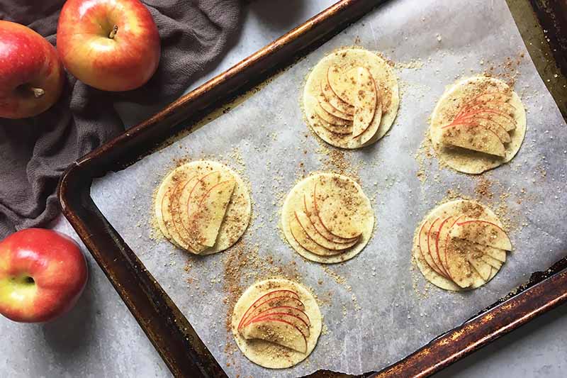 Horizontal image of 6 unbaked apple tartlets on a baking sheet topped with parchment paper.
