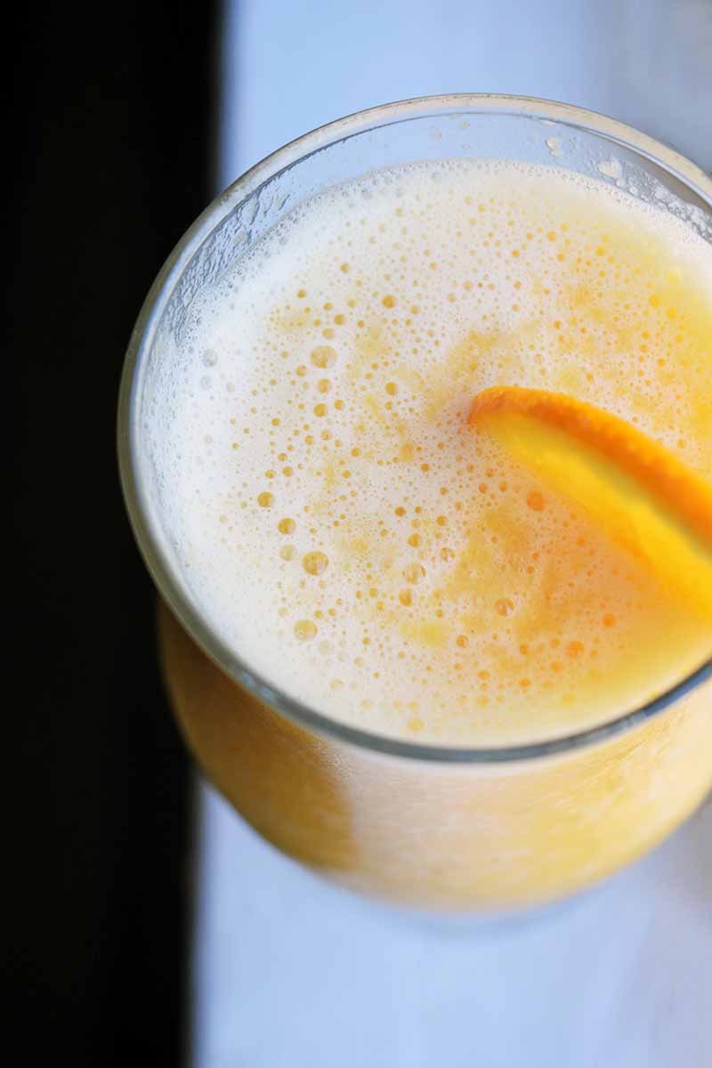 Oblique overhead shot of a smoothie in a glass with a thin slice of orange for garnish, on a white surface with a black background.