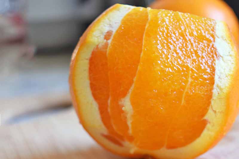 Horizontal image of an orange with about half of the peel sliced away, on a blonde wooden cutting board with more whole fruit in soft focus in the background.