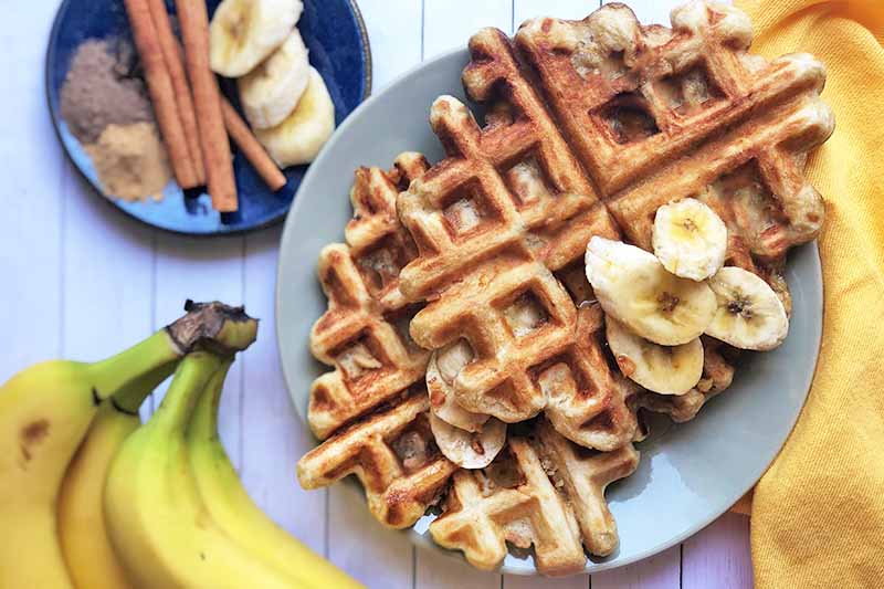 Horizontal image of two golden-brown waffles with bananas on a gray plate, next to a plate of assorted spices.