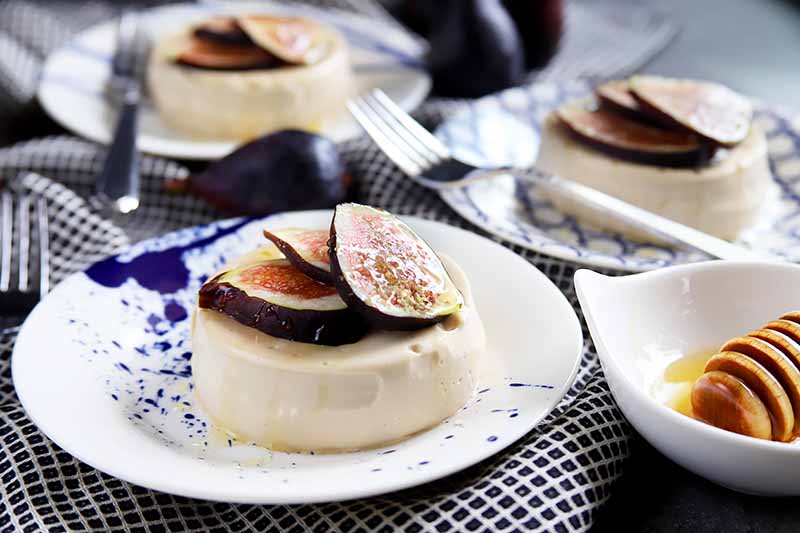 Horizontal image of three plates with panna cotta garnished with figs next to a bowl of honey and metal forks.