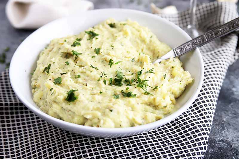 Horizontal image of a white bowl with creamy potatoes with a metal spoon on a checkered towel.