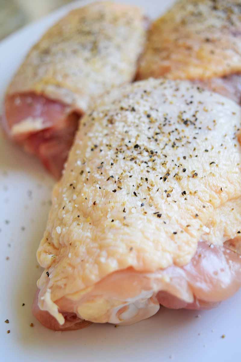 Vertical closeup image of chicken thighs with the skin on, sprinkled with salt and pepper, on a white plastic cutting board.