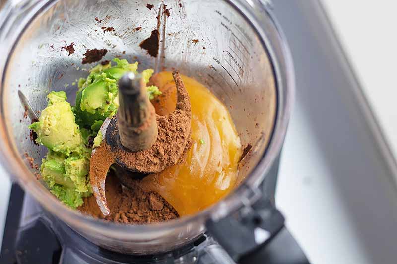 Horizontal image of a food processor with avocado, cocoa powder, and sweetener.