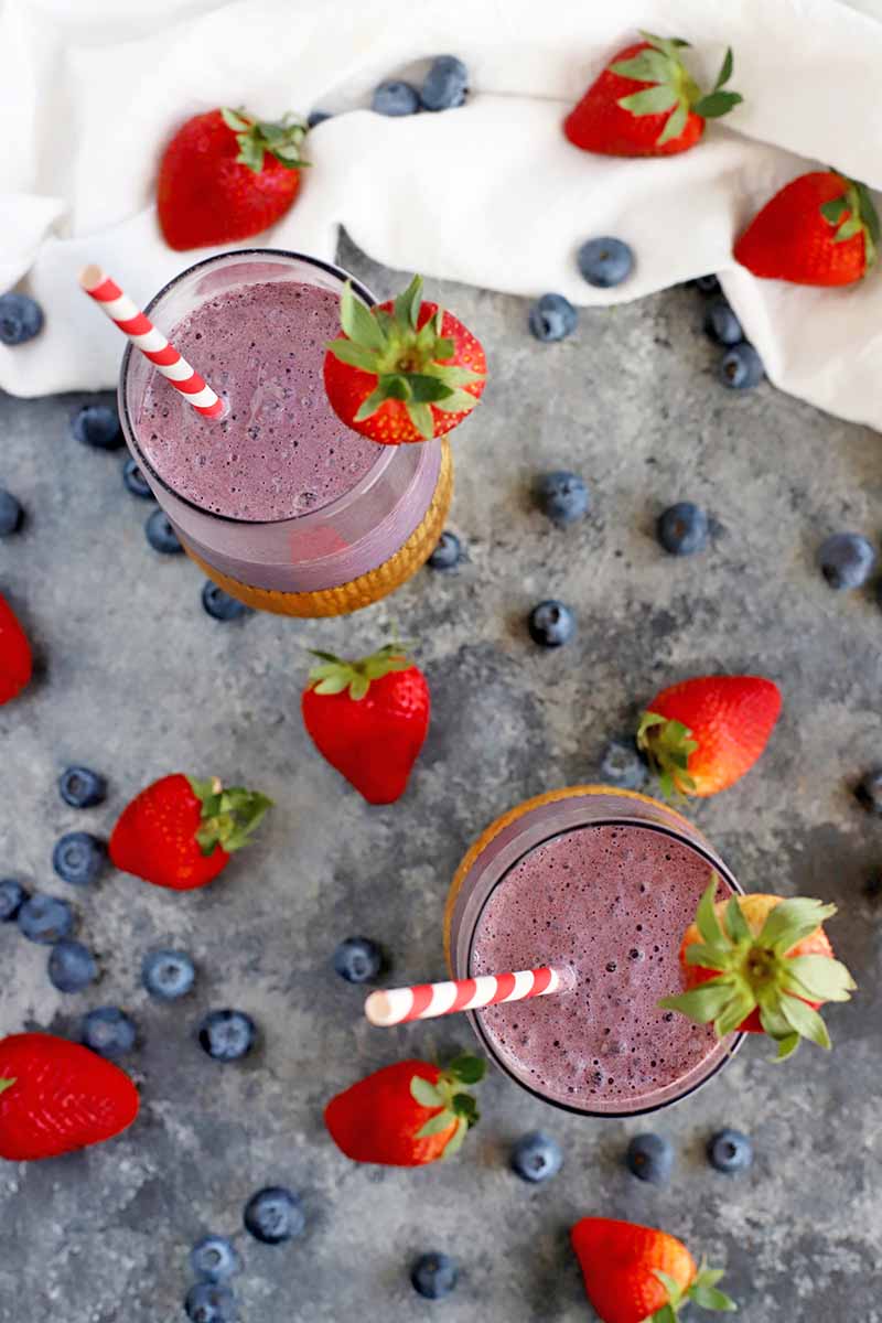 Vertical overhead image of two glasses of purple smoothie, with red and white striped paper straws and berry garnishes, on a gray surface with scattered whole blueberries and strawberries, and a gathered white cloth at the top of the frame.