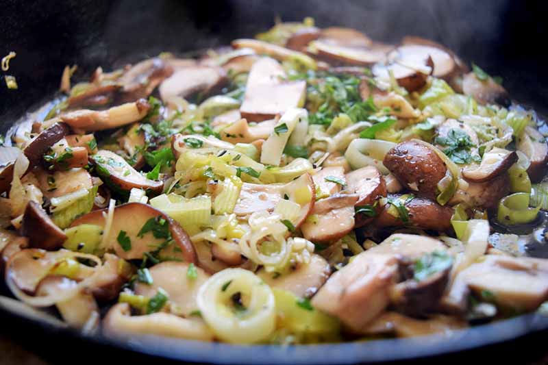 Horizontal closeup image of a cast iron pan of sauteeing sliced mushrooms and leeks.