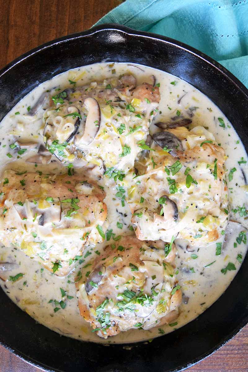 Overhead vertical image of a cast iron pan of chicken fricassee with mushrooms in cream sauce, on a brown wood surface with a blue cloth.