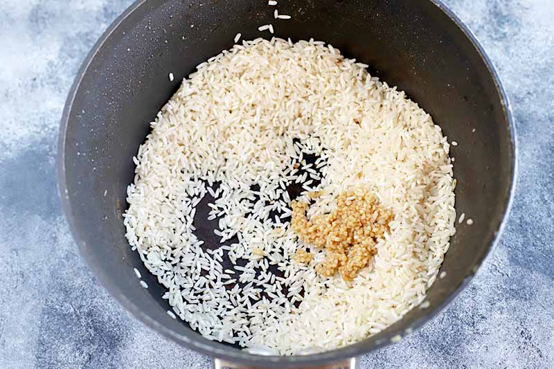 Horizontal overhead image of long grain rice and minced garlic in the bottom of a nonstick cooking pot, on a mottled blue-gray and white surface.