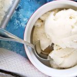 Horizontal overhead image of a white dish of vanilla ice cream with a spoon sticking out of it, with more to the left in a metal loaf pan, and a white kitchen towel topped with a whole pod at the bottom of the frame, on a blue surface with white speckles.