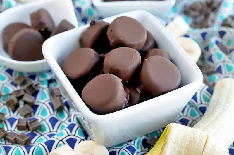 Horizontal image of one medium-sized and two smaller white ceramic dishes of chocolate-covered banana slices, surrounded by more whole and cut fruit and candy chunks on a dark and light blue patterned cloth.