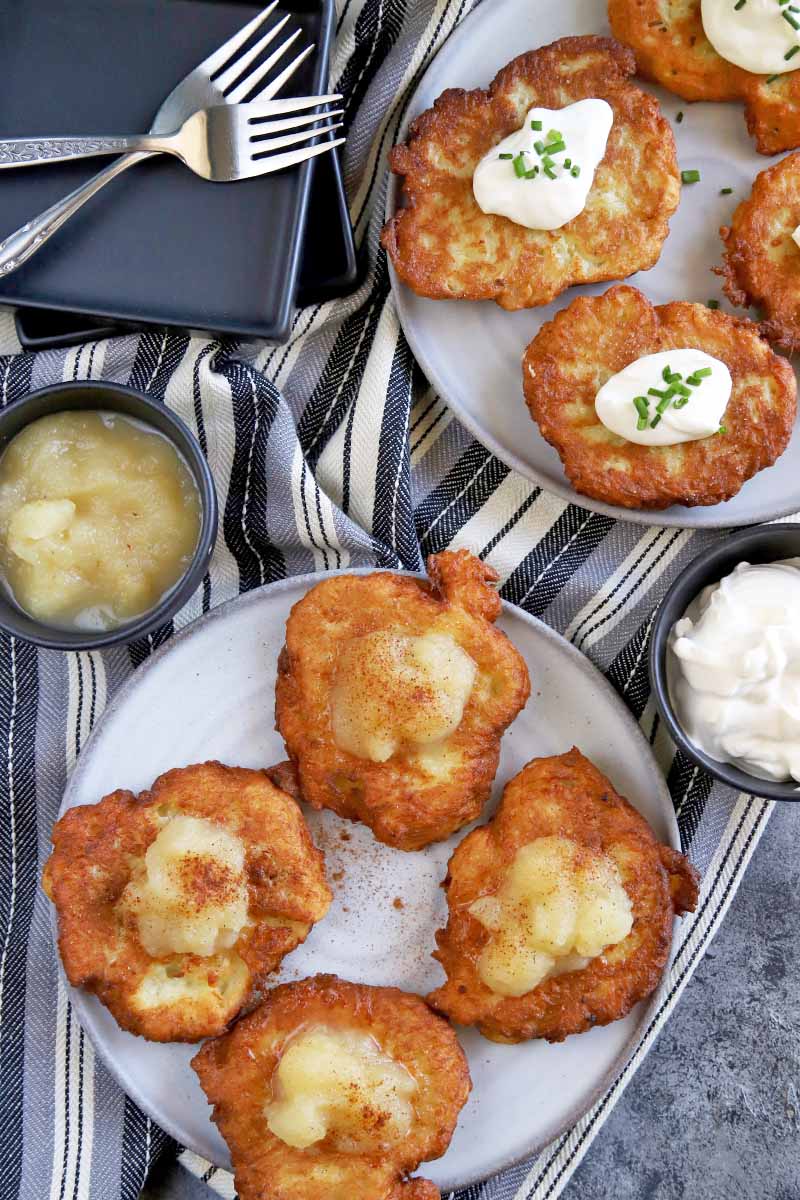 Vertical overhead image of one white plate of four potato pancakes topped with applesauce and cinnamon, and another plate of four kartoffelpuffer topped with sour cream and chives, on a gray surface with a striped black, white, and gray cloth, a short stack of black square dishes and several forks, and small bowls of additional garnishes.