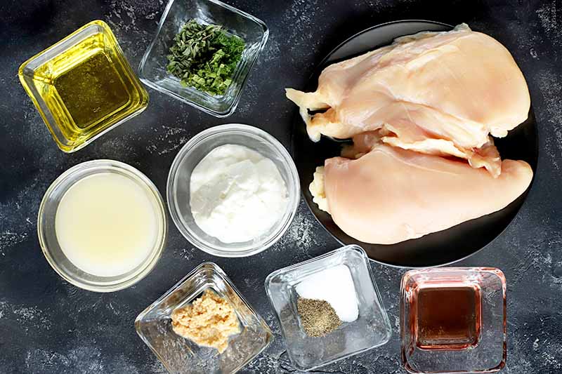 Horizontal overhead image of small round and square clear glass dishes of oil, lemon juice, yogurt, chopped fresh herbs, minced garlic, salt, pepper, and red wine vinegar, with several boneless skinless chicken breasts on a black plate, on a gray surfact.
