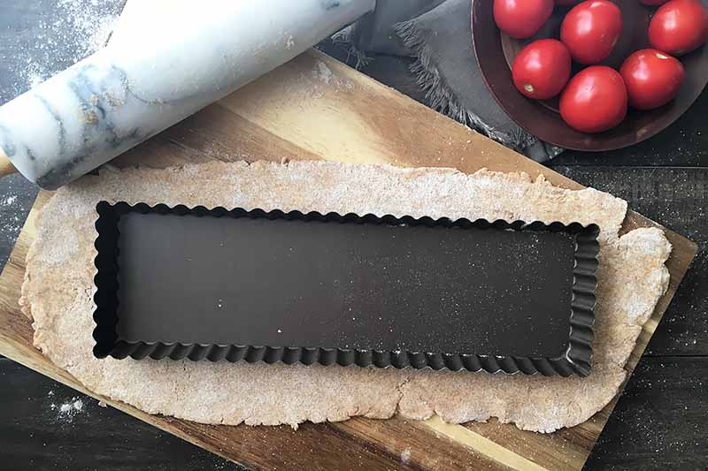Horizontal image of a dark empty tart pan on top of dough. on a wooden surface next to a rolling pin and a plate with fresh produce.