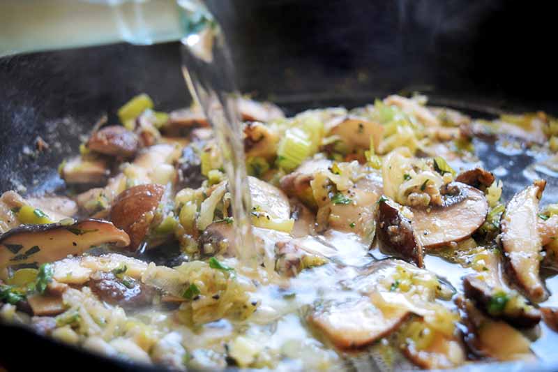 Horizontal closeup image of liquid being poured from a glass bottle into a cast iron pan of sliced mushrooms and sauteed leeks.