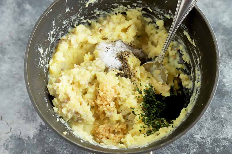 Horizontal image of a pot with creamy potatoes and small piles of salt, garlic, and parsley.