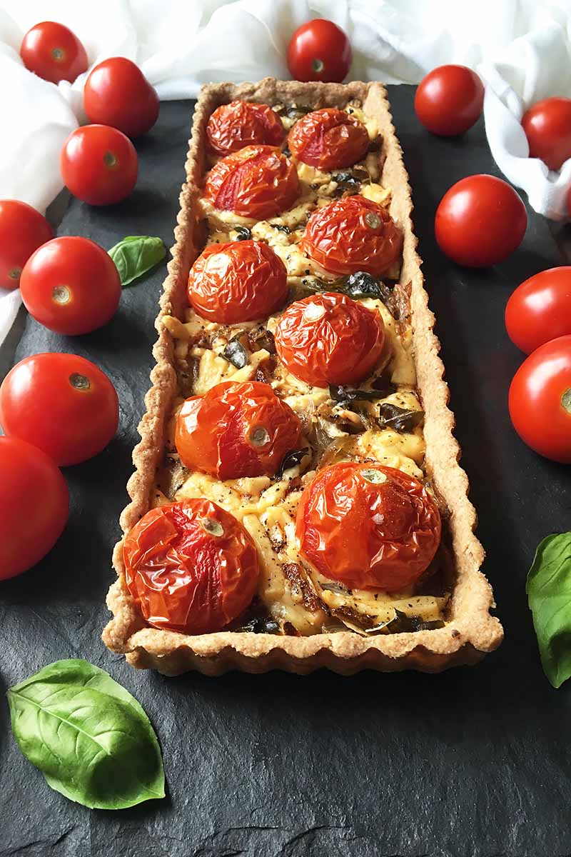 Vertical image of a whole rectangular savory pastry topped with blistered tomatoes and melted cheese on a dark slate.