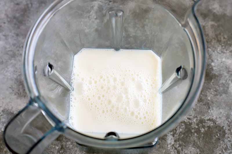 Horizontal oblique overhead image of milk with bubbles on the surface, in a clear plastic blender pitcher on a gray surface.
