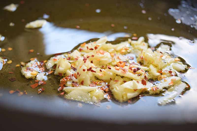 Closeup horizontal image of thinly sliced garlic and red pepper flakes cooking in oil in a frying pan.