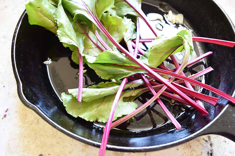 Horizontal image of fresh produce with purple stems in a cast iron skillet with oil and slices of garlic.