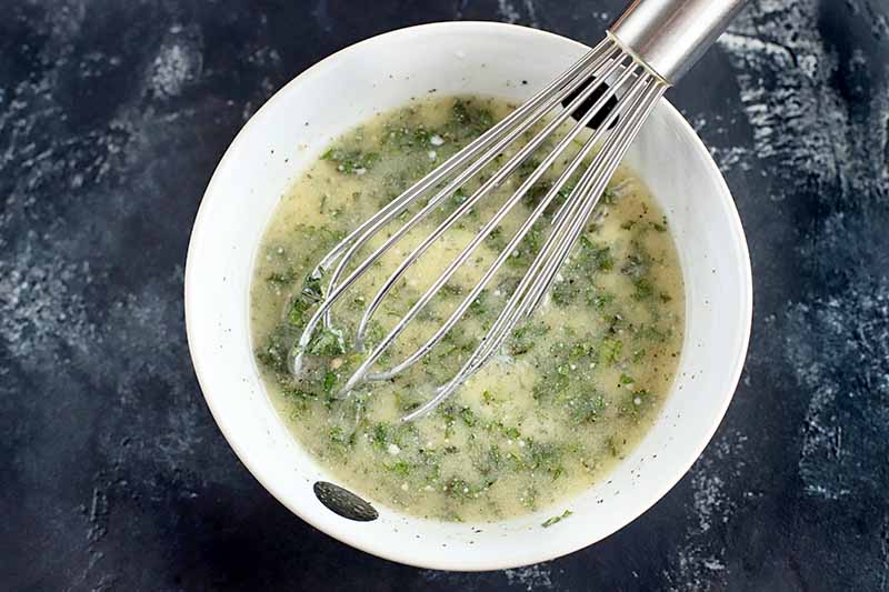 Horizontal image of a pale yellow marinade in a small white creamic bowl, filled with flecks of green fresh herbs, being stirred with a silver wire whisk, on a gray background with white streaks.