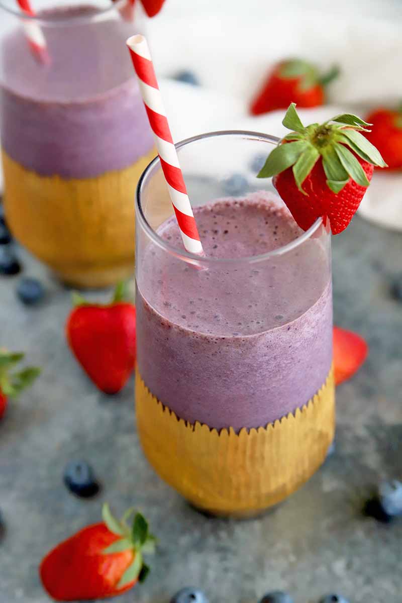 Vertical oblique overhead image of two glasses filled partway with a purple smoothie mixture, wrapped with gold decorative foil wraps at the base of each, with red and white striped purple straws and a berry garnish on the glass in the foreground, on a gray surface with scattered whole blueberries and strawberries, with more nestled in a white cloth in soft focus in the background.