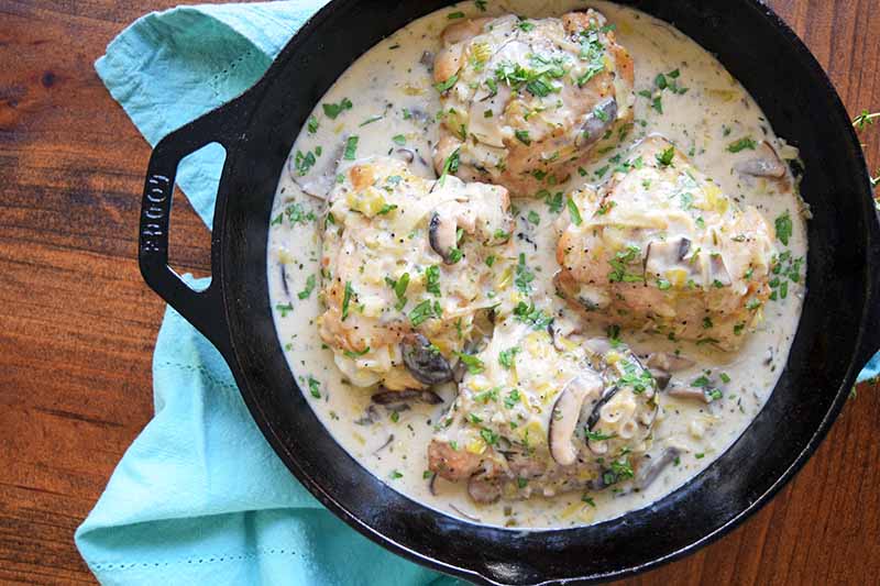 Overhead horizontal image of a cast iron pan of chicken thighs with the skin on, cooked in a cream sauce with leeks and mushrooms, on a wood surface with a gathered light blue cloth dish towel.