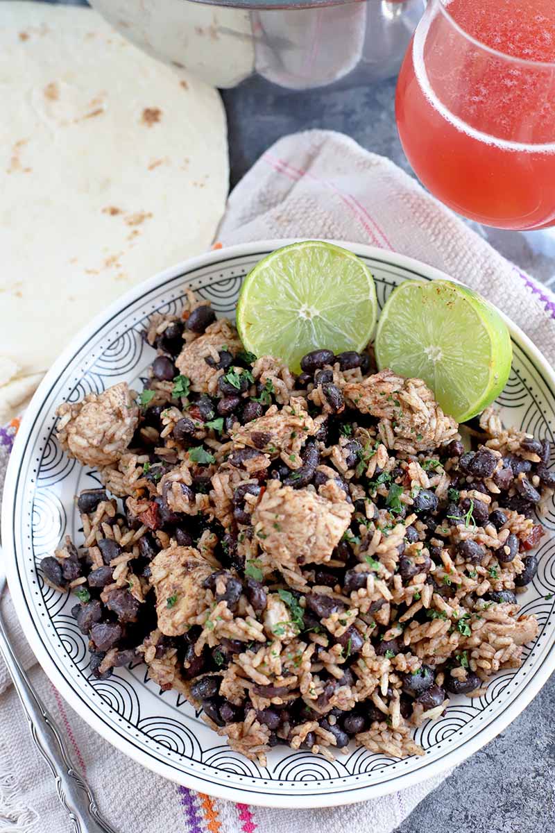 Overhead vertical image of a white plate of chicken with black beans and rice, and two pieces of fresh lime, with tortillas and a wine glass filled with a pinkish red sour beer, on a white cloth with one pink and one orange stripe, on a gray and white surface.