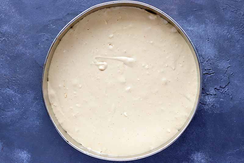 Horizontal overhead image of a metal springform pan filled with a creamy cheesecake filling mixture, on a gray surface with white splotches.