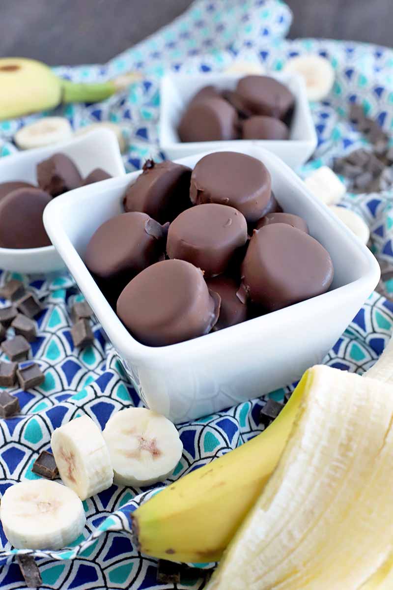 Vertical image of one medium and two small white ceramic bowls of homemade frozen chocolate banana bites, on a dark and light blue patterned cloth with scattered whole, peeled, and sliced fruit, and candy chunks.