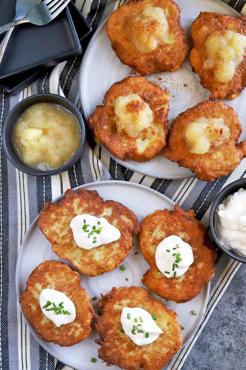 Vertical overhead image of eight potato pancakes arranged on two white plates, half topped with sour cream and chives and the other half topped with applesauce, with two small dishes of these toppings to the left and right, on a striped gray, white, and black cloth with two square black plates and two forks, on a mottled gray surface.