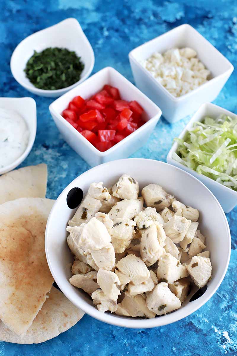 Vertical overhead image of a white bowl of cooked chopped chicken breast in the foreground, surrounded by smaller square and round dishes of chopped tomoat, fresh herbs, feta cheese, shredded lettuce, and yogurt sauce, with several halved pieces of pita bread on top of a blue cloth surface.