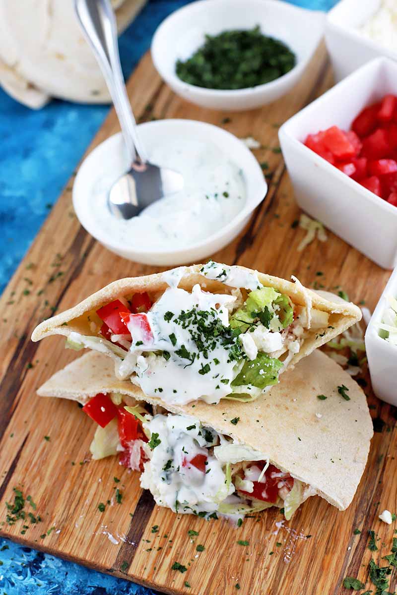 Vertical image of a pita cut in half and filled with chicken, vegetables, yogurt sauce, and fresh chopped herbs, with toppings in square and round white bowls with a spoon stuck into one, on a wooden cutting board on top of a blue cloth, with more bread in the background.