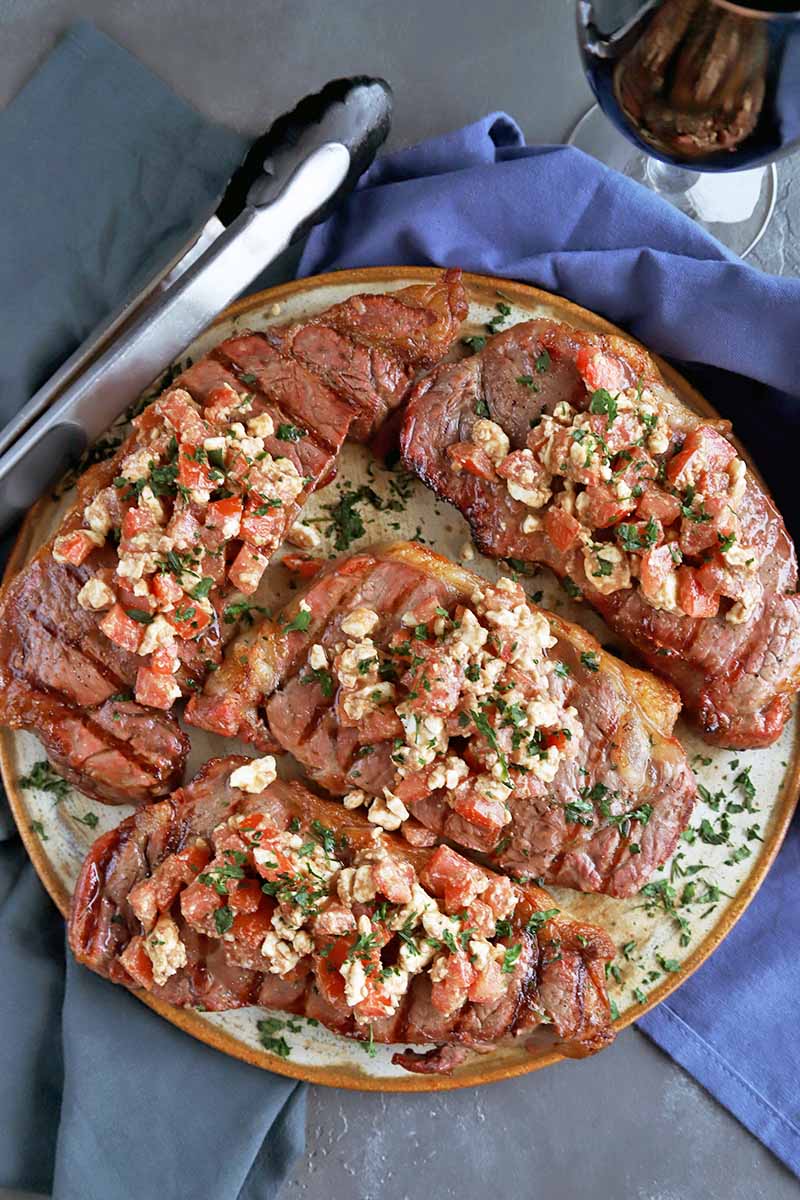 Vertical overhead image of four grilled New York strip steaks topped with herbs, tomato, and feta, on a serving platter with a silver metal and black plastic set of tongs, two blue cloth napkins, and a glass of red wine, on a gray surface.