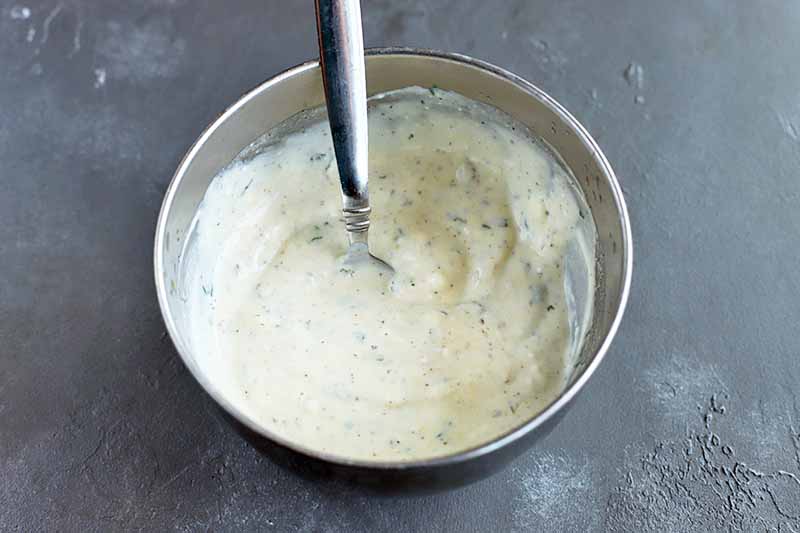 Horizontal overhead image of a small stainless steel bowl of a yogurt-based salad dressing with a spoon for stirring, on a gray background.