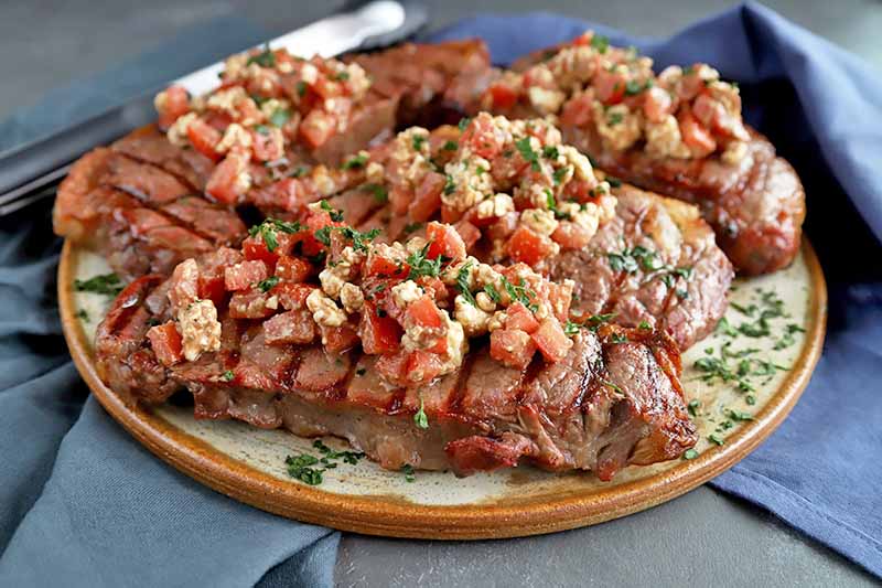 A platter of grilled steak with crumbled feta and tomato salsa, on a gray surface with silverware and two gray-blue cloth napkins.