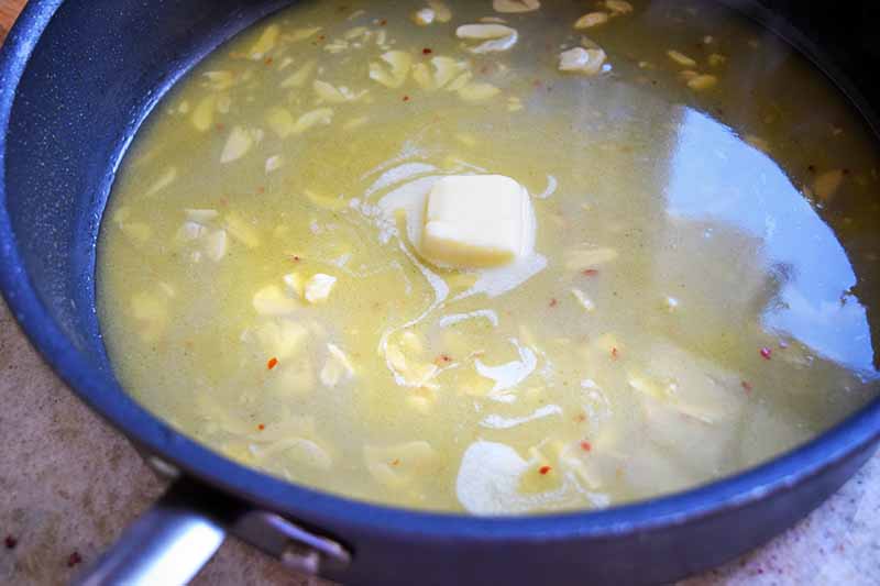 Horizontal image of butter melting in a pan of liquid with thinly sliced garlic and red pepper flakes, in a large frying pan on a beige speckled countertop.