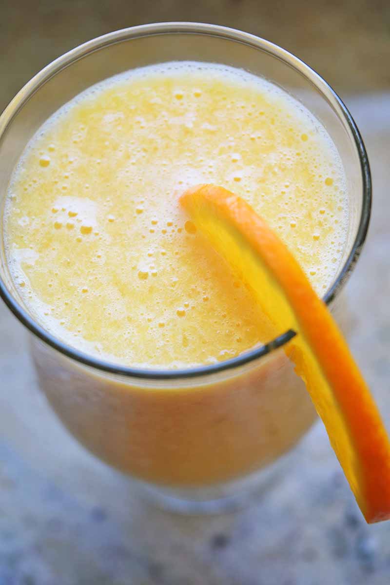 Vertical oblique overhead image of a glass filled with a smoothie and garnished with a fresh orange slice, on a speckled gray surface.
