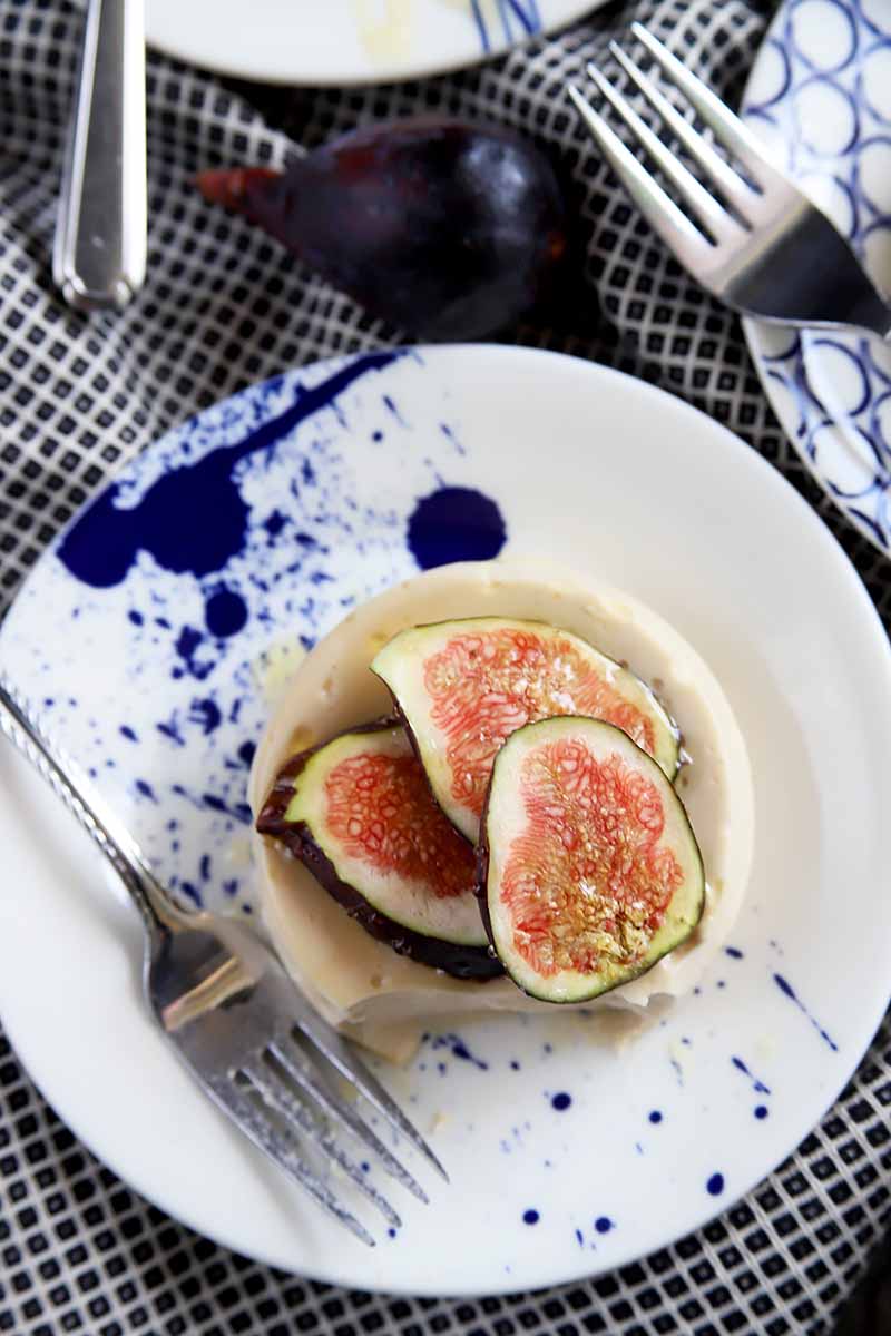 Vertical top-down image of a cream dessert topped with three sliced figs on a blue and white plate next to metal forks.