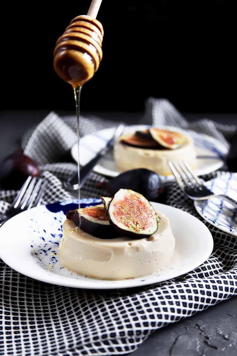 Vertical image of two plates with panna cotta garnished with figs on black and white napkins, with honey pouring over one.