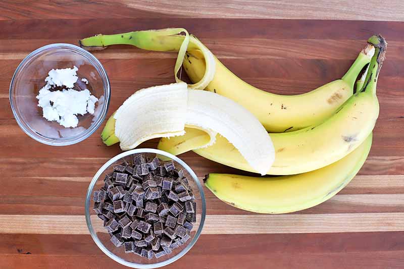 Overhead horizontal image of whole and peeled yellow bananas, and small glass bowls of solid coconut oil and candy chunks, on a striped tan and brown wood surface.