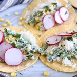 Three yellow corn tortillas topped with a mixture of corn and green beans in a white sauce, garnished with thinly sliced radish, on a speckled gray ceramic plate on top of a striped gray and white cloth, on a brown surface.