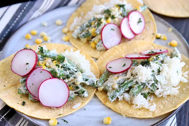 Three yellow corn tortillas topped with a mixture of corn and green beans in a white sauce, garnished with thinly sliced radish, on a speckled gray ceramic plate on top of a striped gray and white cloth, on a brown surface.