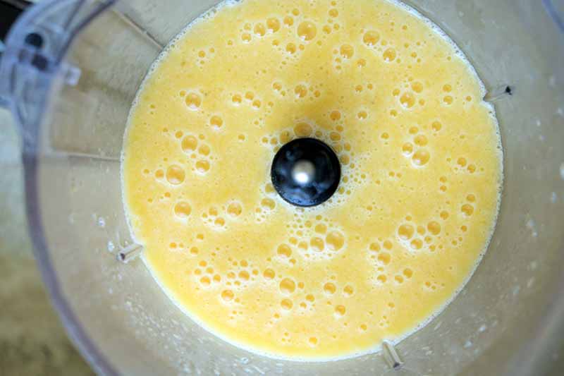 Horizontal closely cropped overhead image of a frothy pale orange smoothie in the bottom of a clear plastic blender canister, on a beige surface.